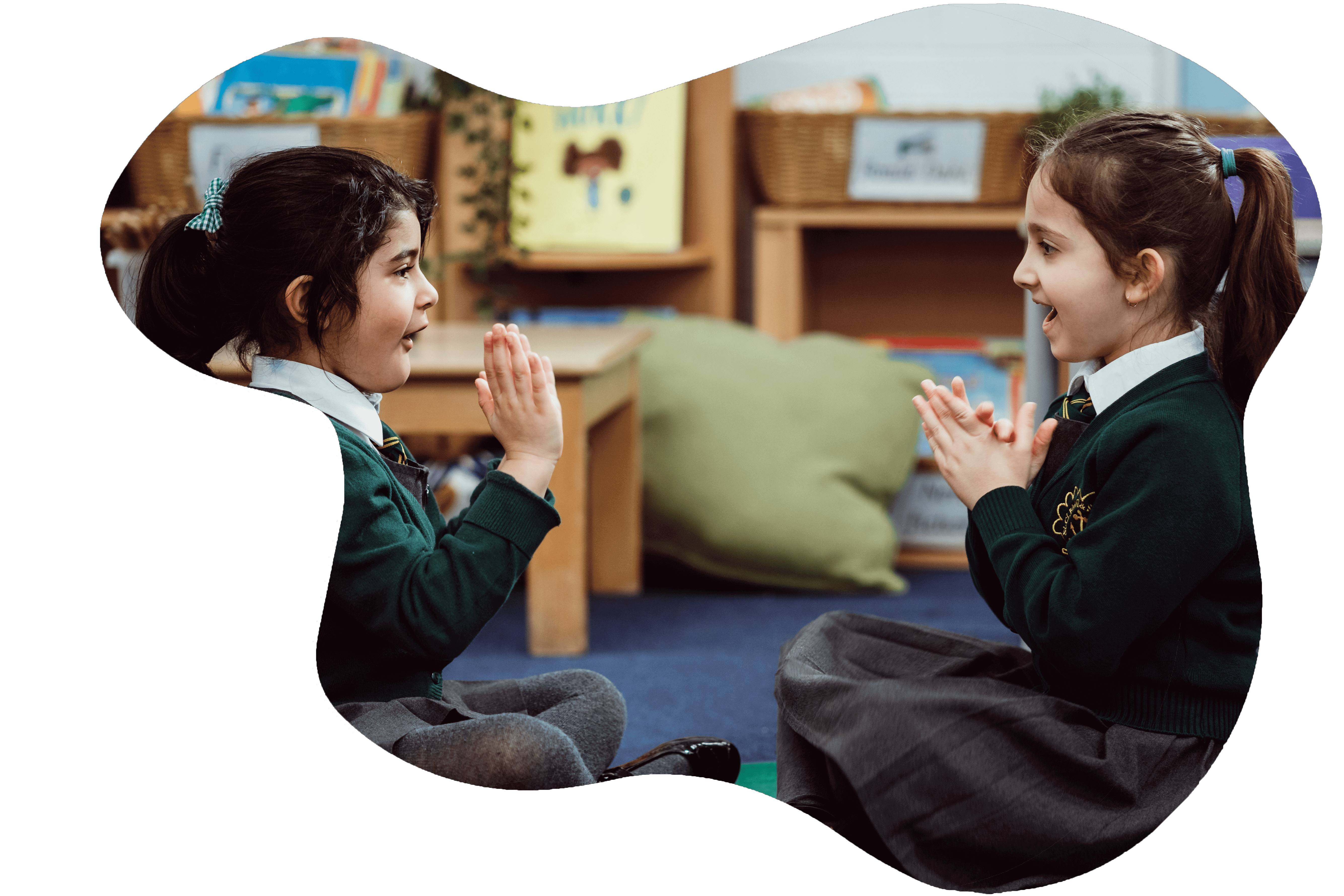 Two young students in school uniforms are seated on the floor facing each other, their hands up as if in a clapping game, sharing a moment of connection and joy in a classroom setting filled with educational materials. Their expressions suggest engagement and happiness in the learning activity they are participating in.