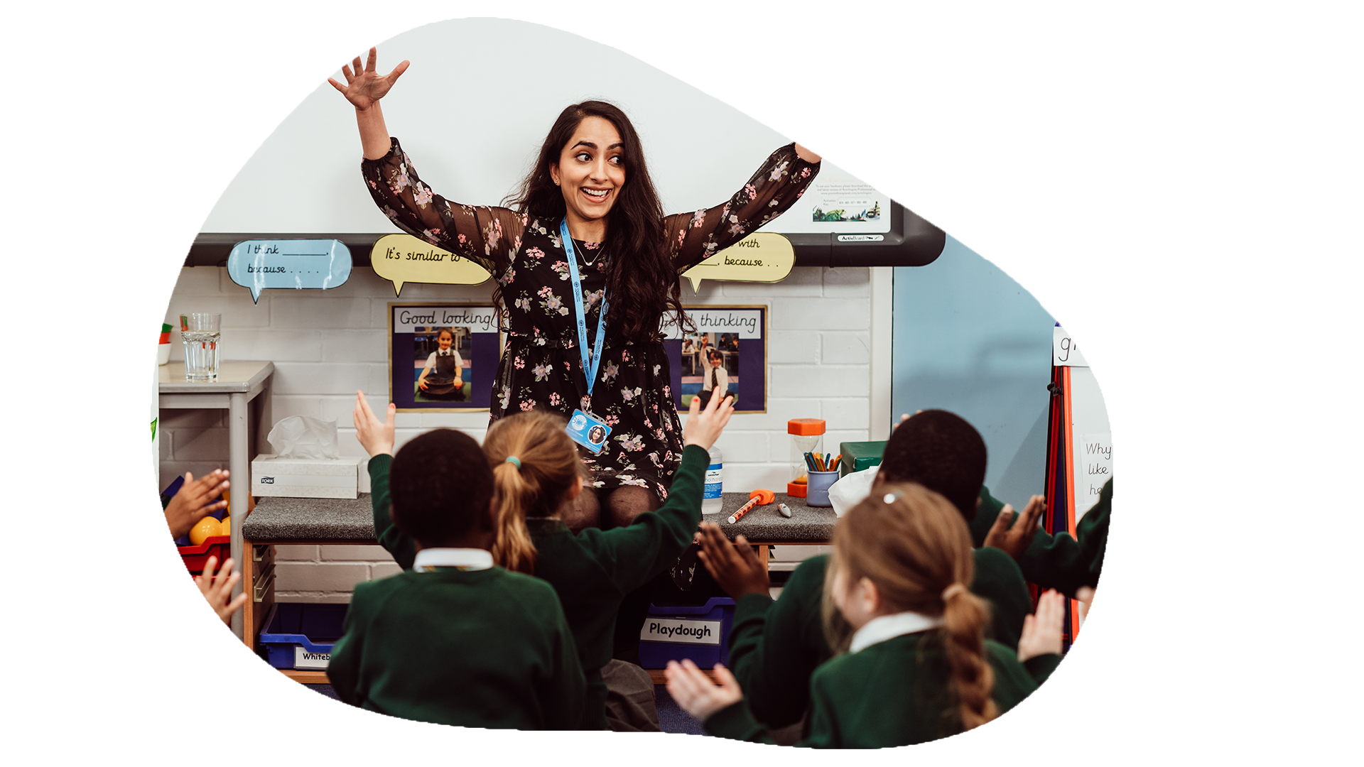 A joyful teacher with outstretched arms stands in front of a classroom, engaging with young students in green uniforms who are enthusiastically raising their hands, creating a lively and interactive learning environment. The classroom is adorned with educational materials, reflecting a focus on creative and active participation.