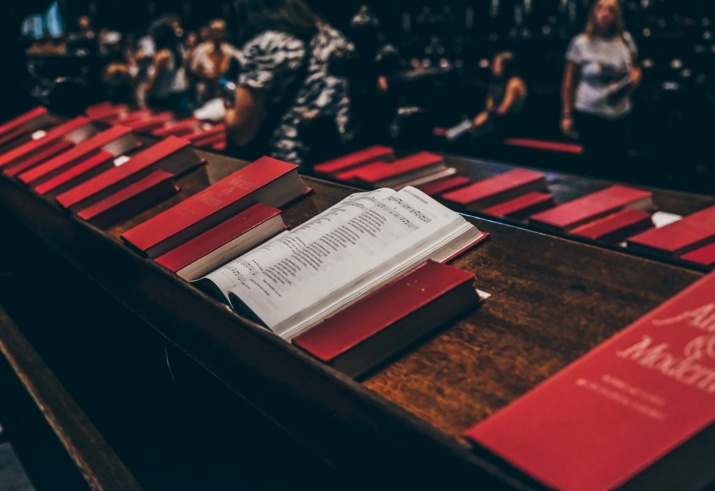 The image shows a row of wooden pews with red hymnals neatly placed on the shelf of each pew. One hymnal is open, displaying sheet music and lyrics, suggesting a setting where congregational singing or a choral performance might take place. In the softly lit background, blurred figures can be seen, adding to the sense of a communal gathering or worship service.