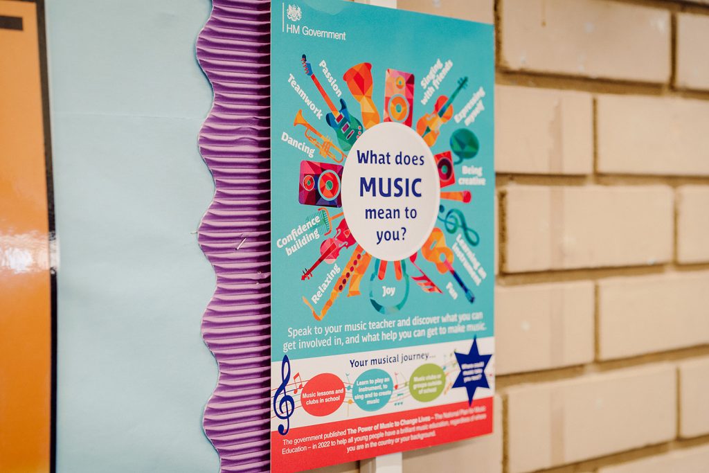 A colorful educational poster is displayed on a brick wall, asking "What does MUSIC mean to you?" with words like "Joy," "Confidence building," and "Teamwork" surrounding vibrant illustrations of musical instruments. The poster encourages dialogue with music teachers and highlights the musical journey, including lessons and clubs in school, with a nod to a government-published report on music's power to change lives.