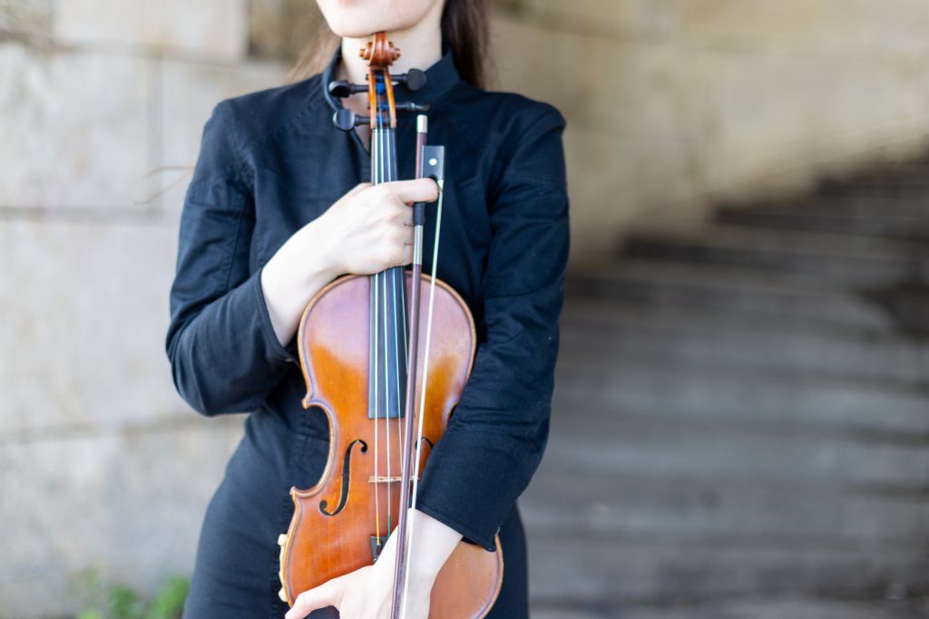 A person in a black outfit holds a violin and bow, ready to play, with a blurred staircase in the background, suggesting an urban or outdoor setting. The focus is on the violin and the musician's hands, conveying a sense of preparation before a performance.