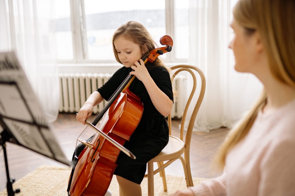 A young girl sits focused on playing a cello, her bow carefully drawing across the strings, while a woman, possibly her instructor, watches attentively from the side. The setting is a bright room with natural light streaming through a window, creating an atmosphere of a calm and dedicated music lesson.