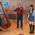 A group of five adults are engaged in a lively music education session in a room with wooden paneling. A woman plays a double bass, another holds a blue balloon aloft, while the others watch with hands raised in anticipation, surrounded by colorful markers on the floor, creating an atmosphere of interactive learning and enjoyment.