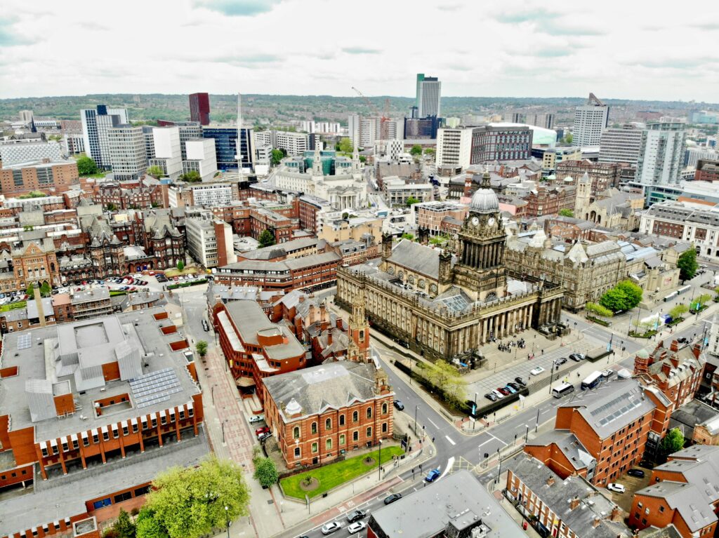 A photo taken from above showing a view of Leeds. In the background you can see trees and landscapes and in the foreground there are a huge mixture of old buildings, skyscrapers and apartments.